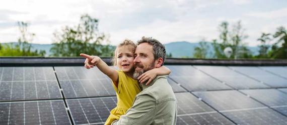 Mann mit Kind auf dem Arm vor PV-Fläche.