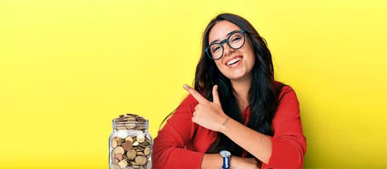 Dunkelhaarige junge Frau mit Brille zeigt lachend nach links oben. Neben ihr steht ein Glas mit Geldmünzen