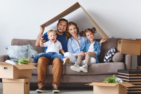 Eltern mit Kleinkindern sitzen auf dem Sofa und halten einen Karton als Hausdach über sich