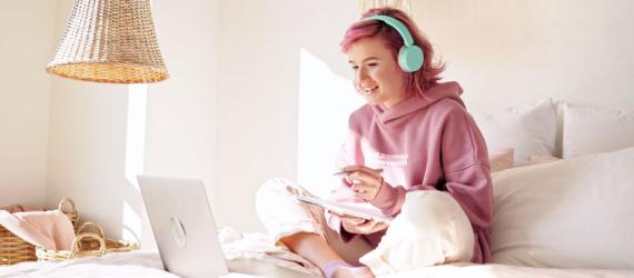 Junge Frau mit Kopfhörern sitzt auf dem Bett vor ihrem Laptop