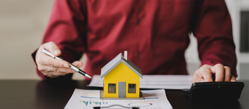 In Immobilien investieren: Mann zeigt mit Kugelschreiber auf Modellhaus auf Schreibtisch