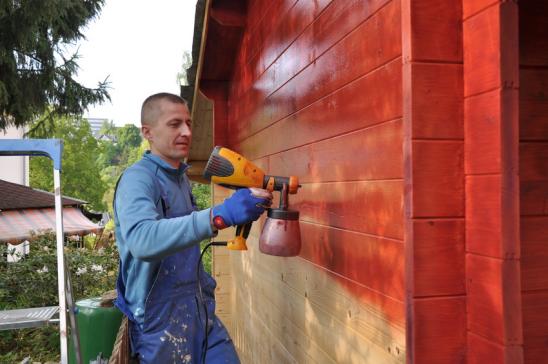 Mann sprüht roten Holzschutz auf Gartenhauswand