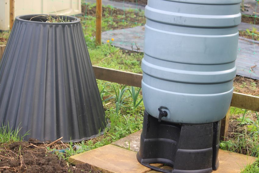 Photo showing a tall grey plastic water butt container (polypropylene) on a black stand, with a tap to fill up watering cans or connect to a hose pipe. The water butt is located on a patio of paving slabs, within a shared allotment vegetable garden. A cone-shaped plastic compost bin / compost heap is situated next to the water butt, being filled with composting garden waste and weeds.
